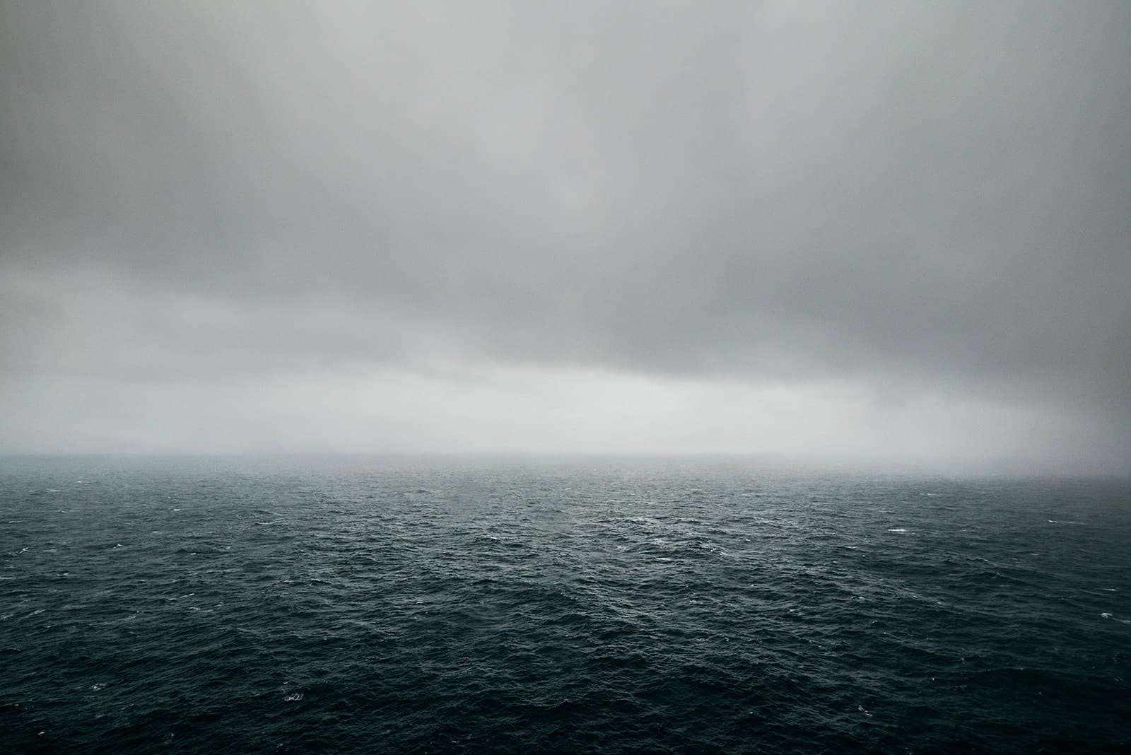 A vast, dark ocean stretches under a heavy, gray sky with fog obscuring the horizon, taken on the Faroe Islands.