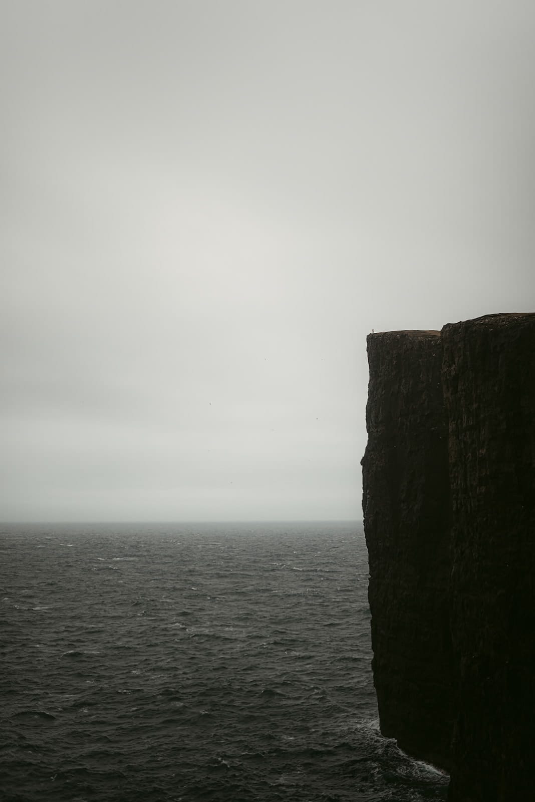 A towering dark cliff rises sharply from a choppy sea under a pale, overcast sky, with a tiny figure standing at the cliff’s edge, taken on the Faroe Islands.