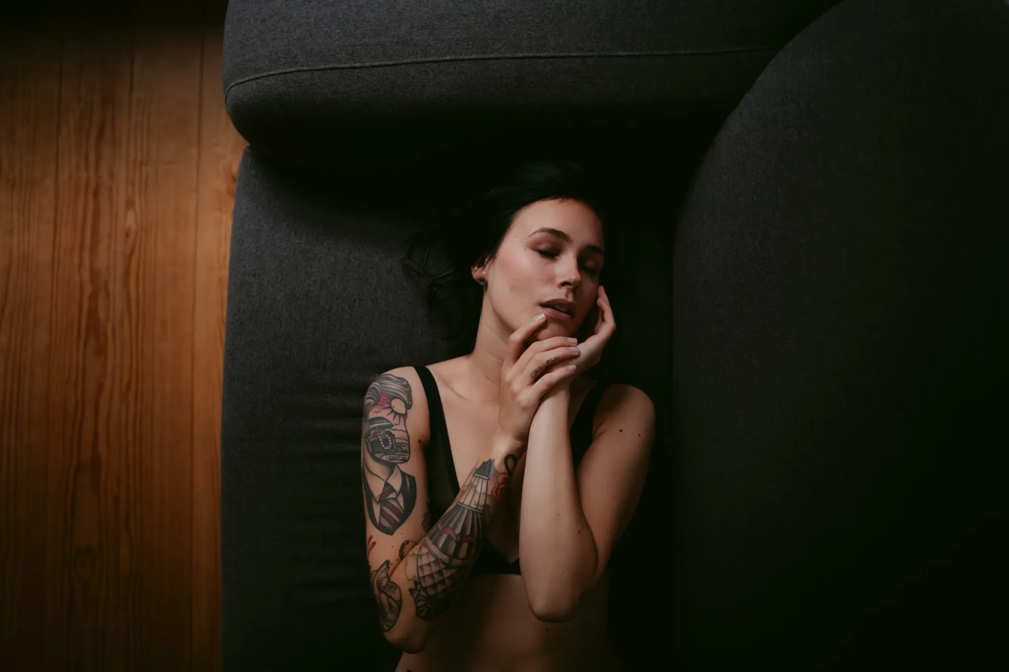 A softly lit portrait of a woman in black lingerie lying on a gray couch with eyes closed and hands to their face, showcasing a colorful sleeve tattoo, taken by Boudoir Photographer Chris Zielecki