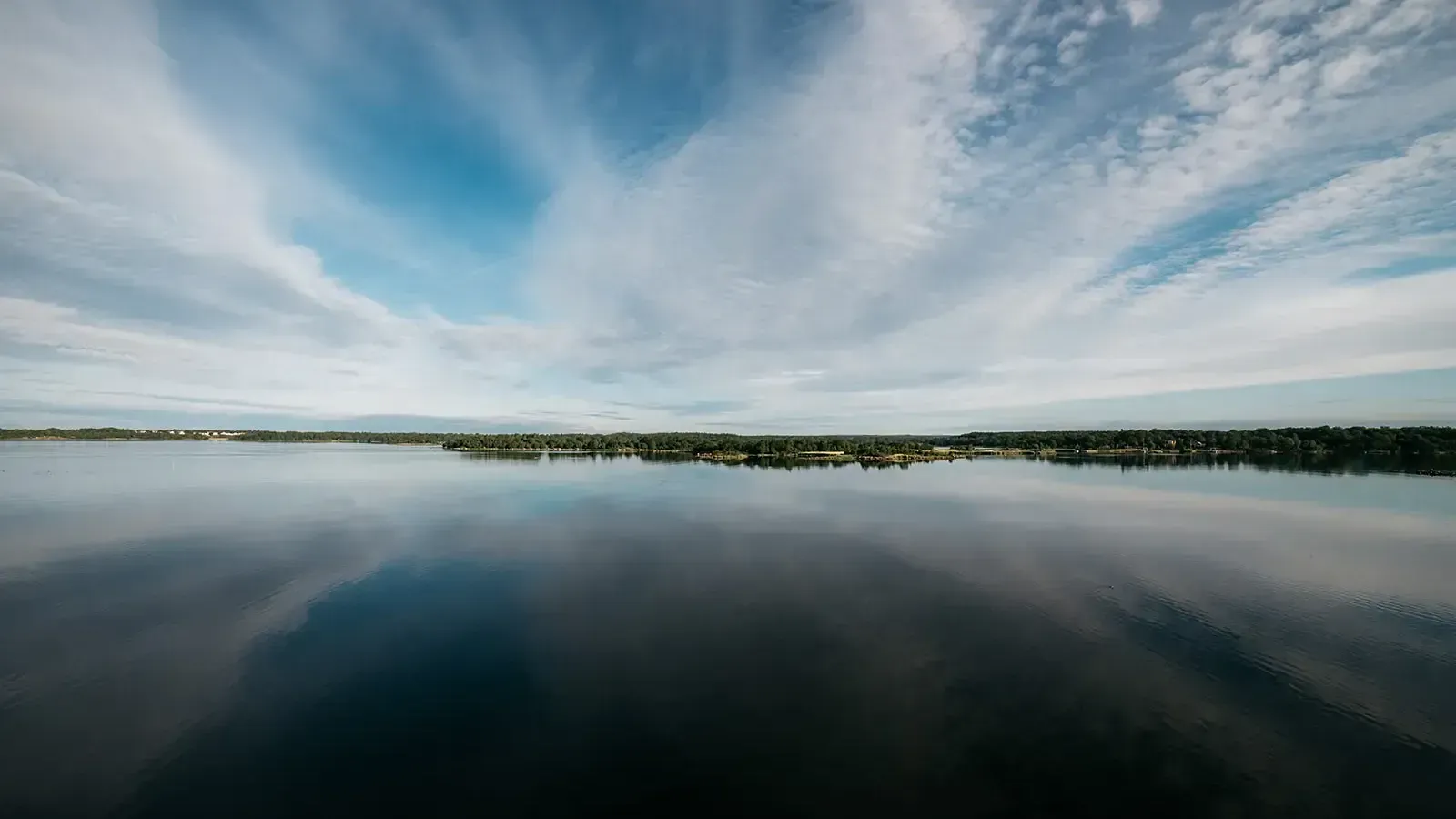 A vast, calm lake reflecting a partly cloudy sky with wispy clouds stretching across the horizon, bordered by a distant tree-lined shore, taken in Sweden by Chris Zielecki