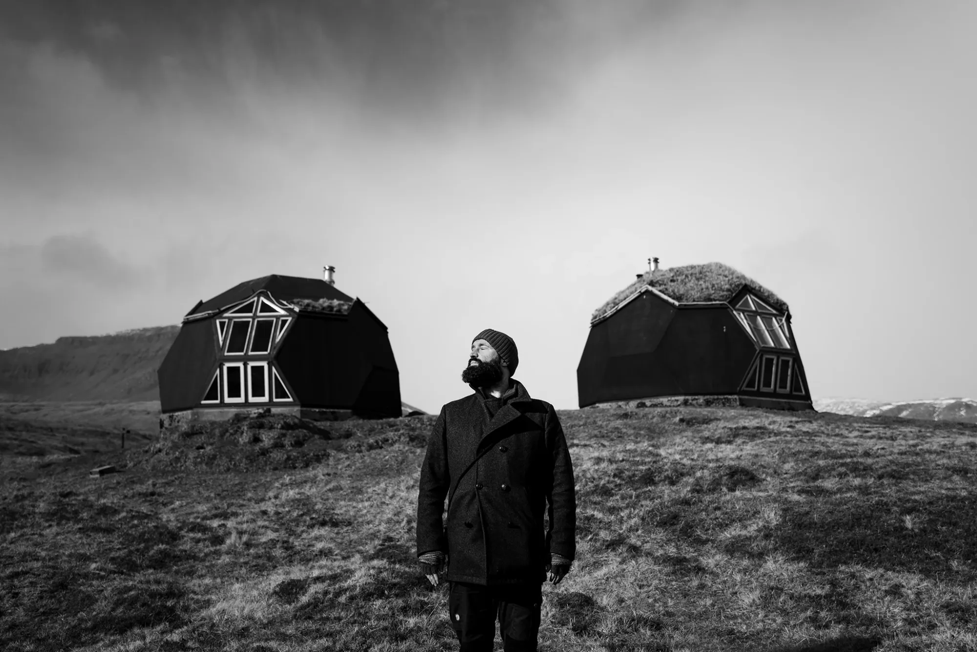 Chris on the Faroe Island Photo Tour wearing a beanie standing in a brown grassy field with a camera hanging from his coat, with two dome-shaped grass-roofed buildings behind him and mountains visible in the distance under an overcast sky