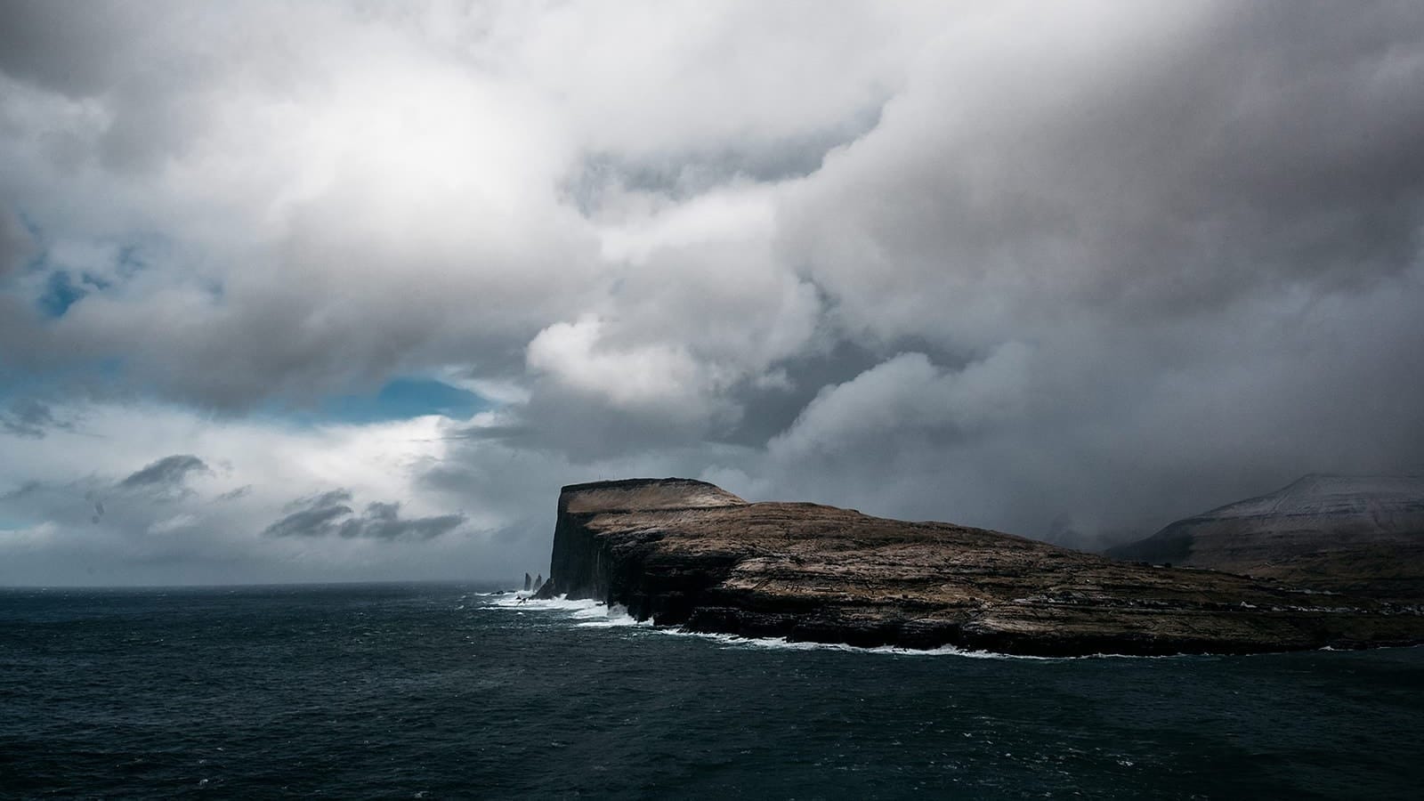 Waves crash against a rugged cliff jutting into a dark sea beneath a dramatic sky of heavy clouds and patches of blue, taken on the Faroe Islands.