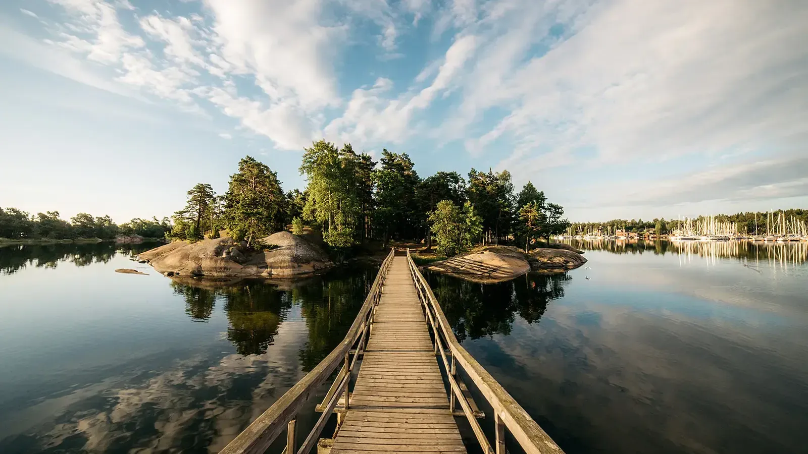 A wooden footbridge stretches across calm water towards a small rocky island, with a marina of sailboats visible in the distance under a partly cloudy sky, taken in Sweden by Chris Zielecki.