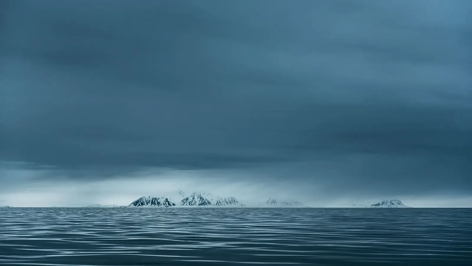 Calm Arctic sea with gentle ripples under a moody, overcast sky, with snow-covered mountains visible on the horizon, photographed in Svalbard by Chris Zielecki
