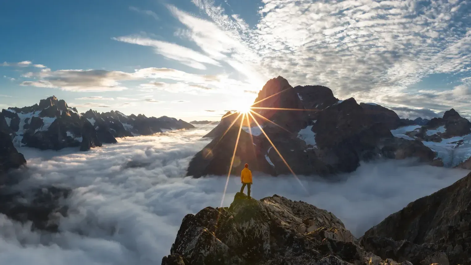 Chris in a yellow jacket stands on a rocky peak overlooking a sea of clouds with jagged, snow-capped mountains bathed in the golden light of the setting sun, captured in Greenland